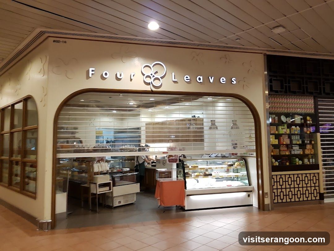 A wide shot of a bakery storefront with a large arched entrance. The sign above reads 'Four Leaves' with a logo of four circles. Inside, there are glass display cases with baked goods, a counter, and shelves of packaged products on the right.