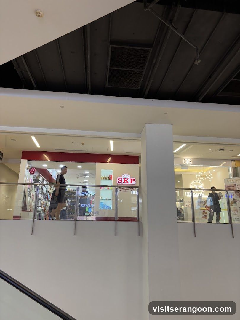A view of a retail store located on an upper floor of a shopping mall. The store has a red and white facade with a prominent illuminated sign reading 'SKP'. A glass railing separates the walkway from the store entrance, and a large white structural column is visible in the foreground.