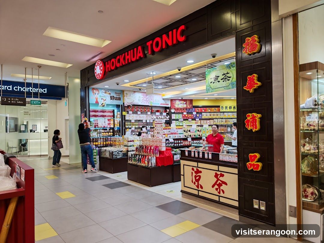 A brightly lit storefront for 'Hockhua Tonic' located within a shopping mall. The shop features a dark brown facade with large red neon lettering for the name and vertical Chinese characters on the right pillar. Inside, shelves are stocked with various packaged goods, and a counter displays more products. A staff member in a red shirt stands behind the counter, and customers are browsing near the entrance.