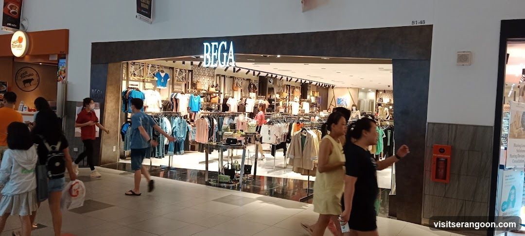 A wide shot of a retail store entrance inside a shopping mall. The store has a large, dark grey facade with the name 'BEGA' in large, illuminated white letters above the opening. Inside, racks of colorful clothing are visible. Several shoppers are walking past the store in the foreground.
