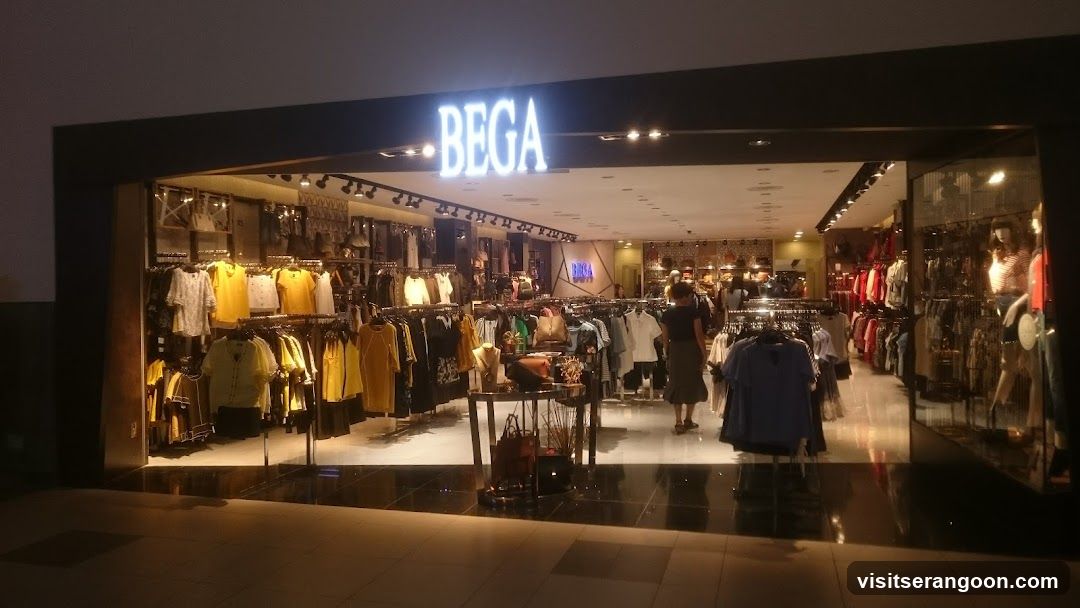 A brightly lit storefront with the illuminated sign 'BEGA' above the entrance. The interior displays racks of clothing, including yellow and black garments, and mannequins wearing outfits. A customer is visible browsing inside.
