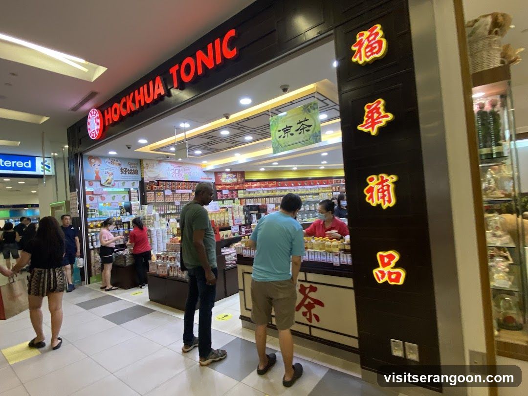 A brightly lit storefront with a large red sign reading 'HOCKHUA TONIC' and vertical Chinese characters on a dark pillar. Several customers are standing at the counter interacting with staff, while others browse shelves of bottled products in the background.