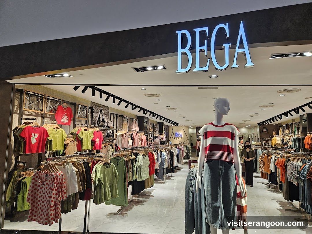 A brightly lit storefront with the name BEGA in large, illuminated blue letters above the entrance. The interior displays racks of colorful clothing, including t-shirts and dresses, alongside a mannequin wearing a striped top and denim skirt.