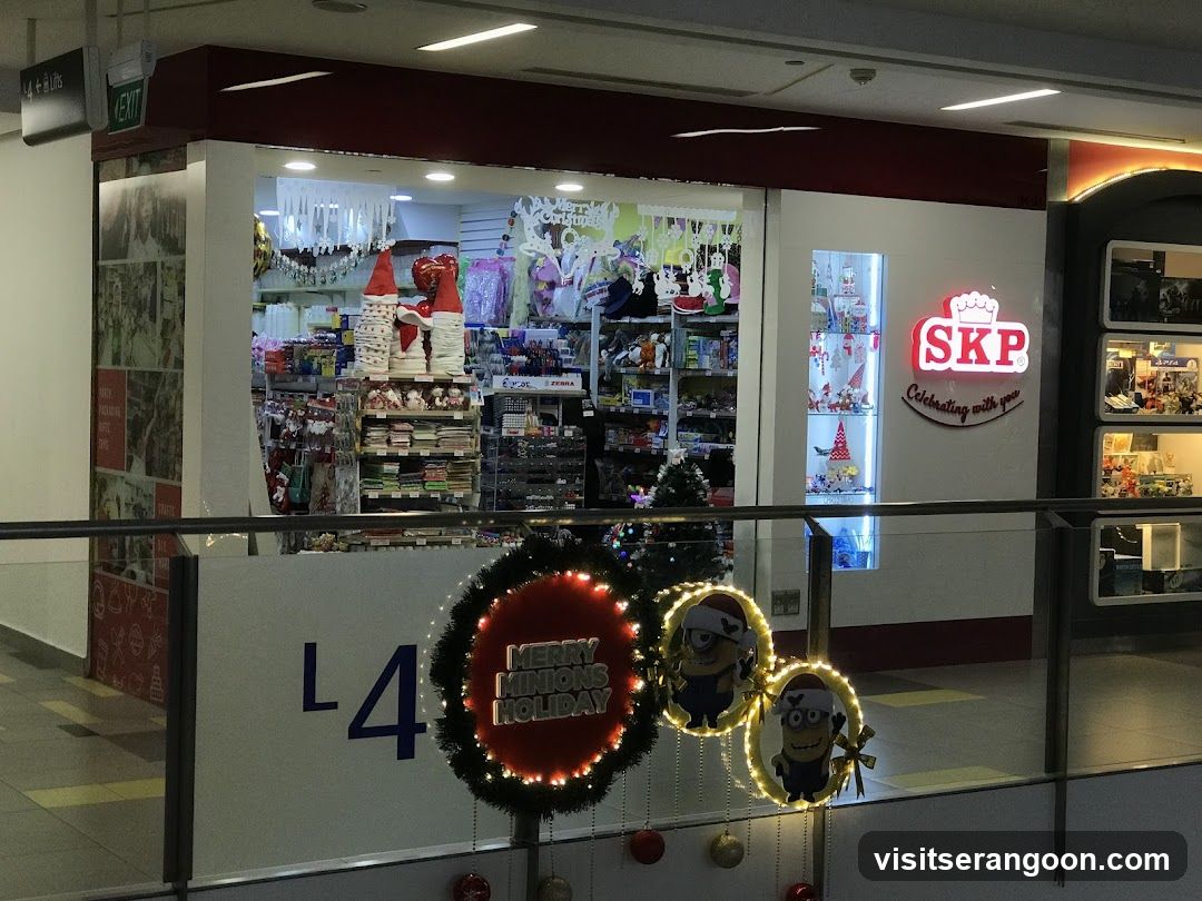 A view of a retail store entrance located on the L4 level of a shopping mall. The storefront features a large glass window displaying shelves of merchandise, including Santa Claus figures and hanging ornaments. To the right of the window is a lit sign reading 'SKP Celebrating with you'. In the foreground, a glass railing is decorated with festive items, including a large red wreath with 'MERRY MINIONS HOLIDAY' text and two smaller wreaths featuring Minion characters.