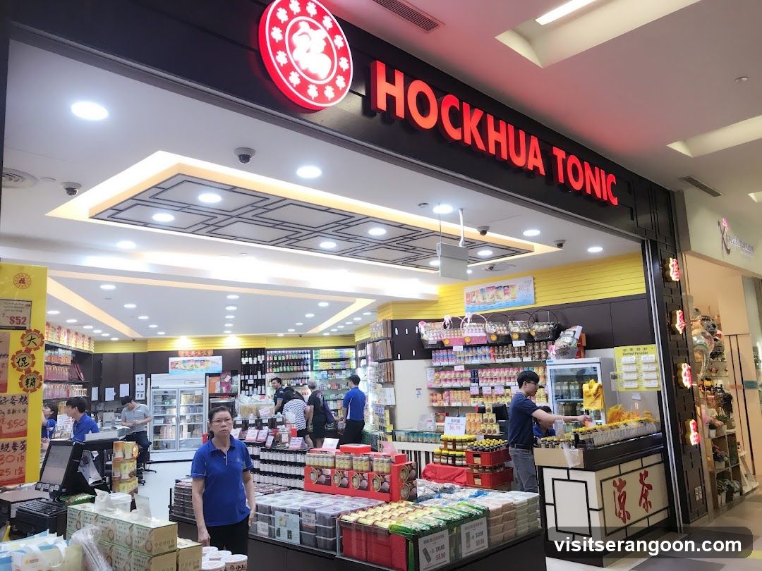A brightly lit storefront with a black facade and a large red sign reading 'HOCKHUA TONIC' in capital letters. A circular red logo with Chinese characters is visible to the left of the text. The store interior is visible through the wide entrance, showing yellow walls, shelves stocked with various bottled goods, and several customers browsing. Staff members wearing blue polo shirts are present inside the store.