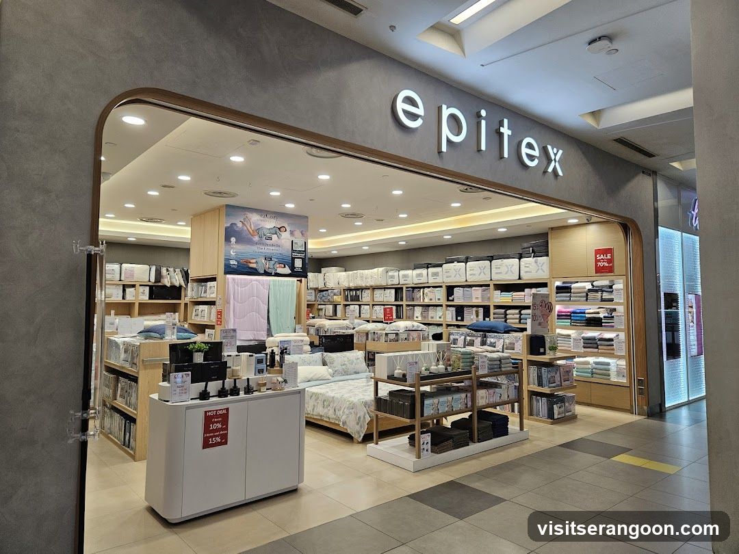 A brightly lit retail store entrance with a large arched opening. The store name 'epitex' is displayed in large, white, illuminated letters above the entrance. Inside, shelves are stocked with bedding and home goods, and a white counter is visible in the foreground.