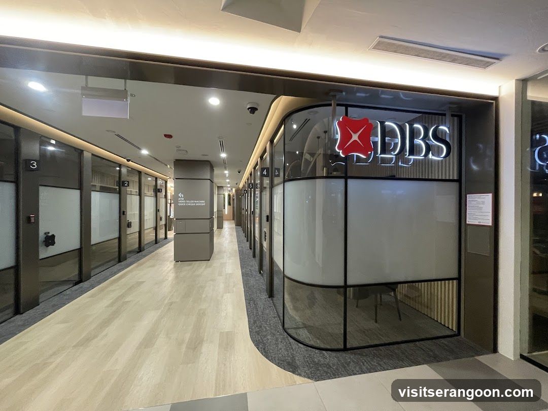 A modern office hallway featuring a glass-walled office on the right with a prominent illuminated DBS logo. The corridor has light wood flooring, dark grey carpeting near the office, and a row of numbered glass doors on the left.