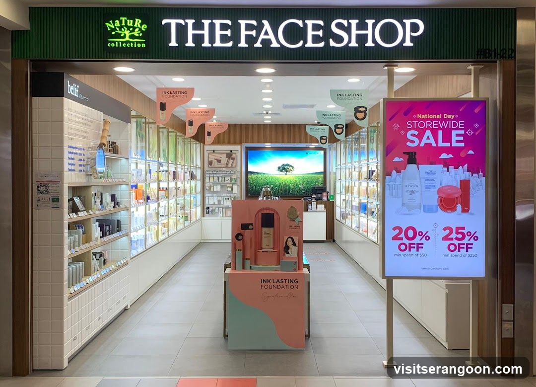 A brightly lit retail store entrance with a green overhead sign reading 'THE FACE SHOP' and 'Nature collection'. The interior is visible, featuring white shelving units stocked with various cosmetic products. A central display stand promotes 'INK LASTING FOUNDATION', and a large digital poster on the right advertises a 'National Day STOREWIDE SALE' with 20% and 25% off discounts.