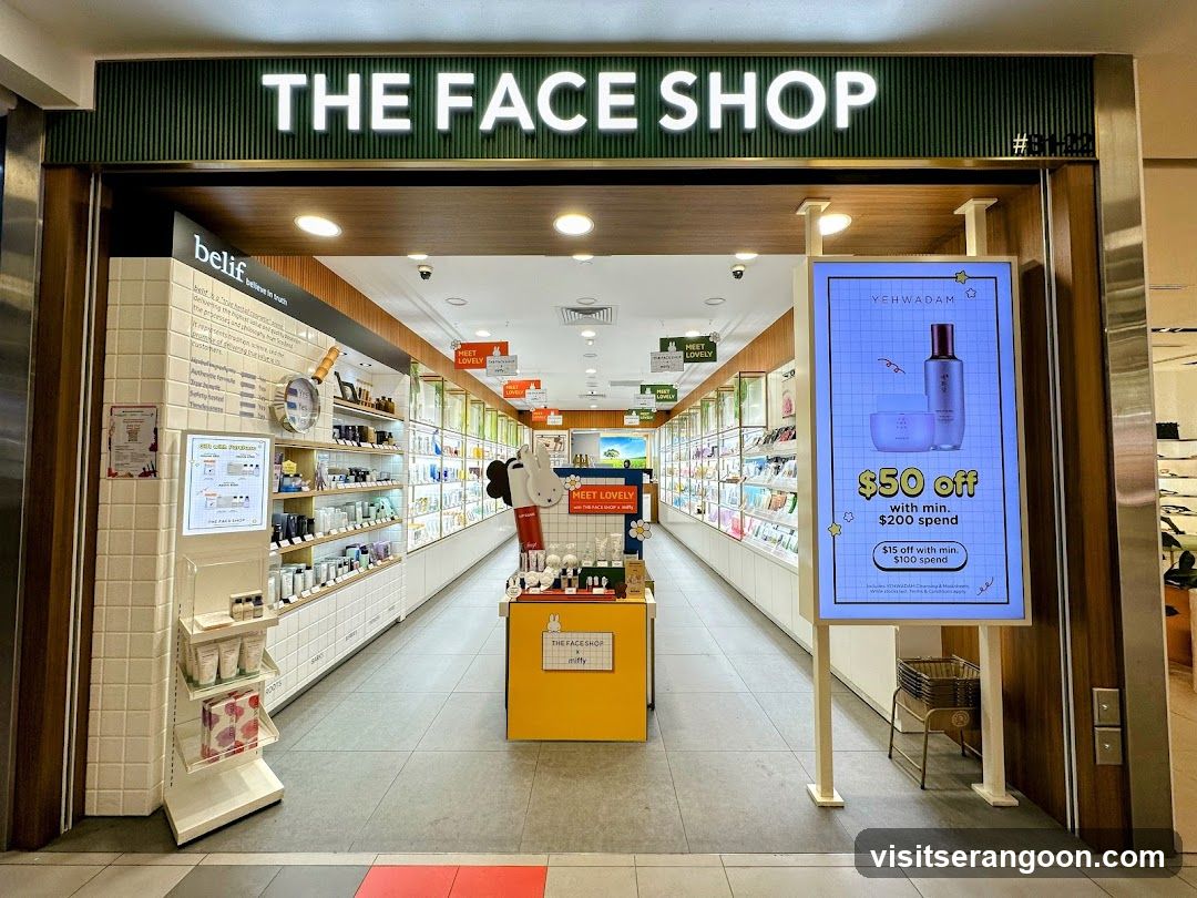 A brightly lit retail store entrance with a large, illuminated sign reading 'THE FACE SHOP' above the doorway. The interior is visible, showing aisles of shelves stocked with beauty products, promotional displays, and hanging signs. A large digital advertisement stand is positioned to the right of the entrance.