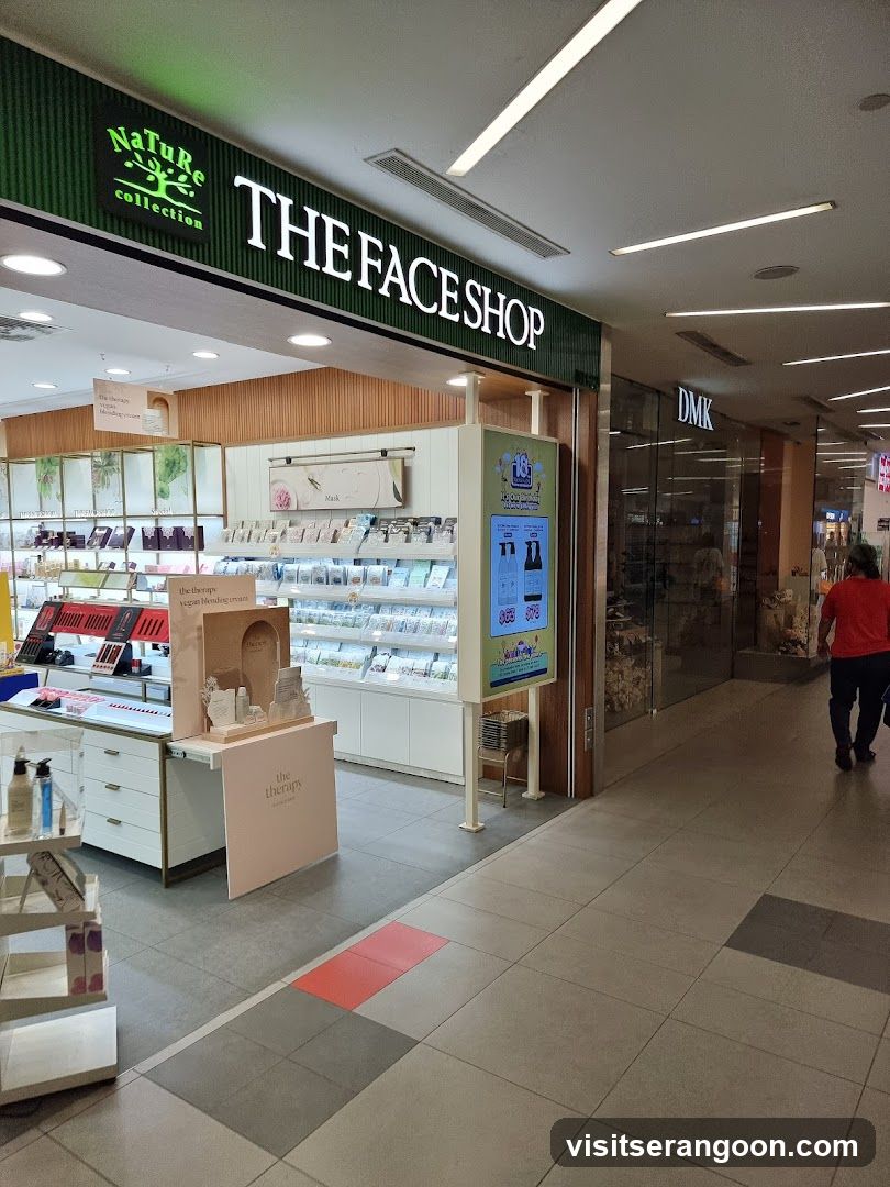 A brightly lit retail storefront for 'THE FACE SHOP' located inside a shopping mall. The store features a green corrugated sign with white lettering and a 'Nature collection' logo. Inside, white shelving units display various cosmetic products, and a promotional stand is visible near the entrance. A digital advertising kiosk stands outside the store, and another store labeled 'DMK' is visible further down the hallway.