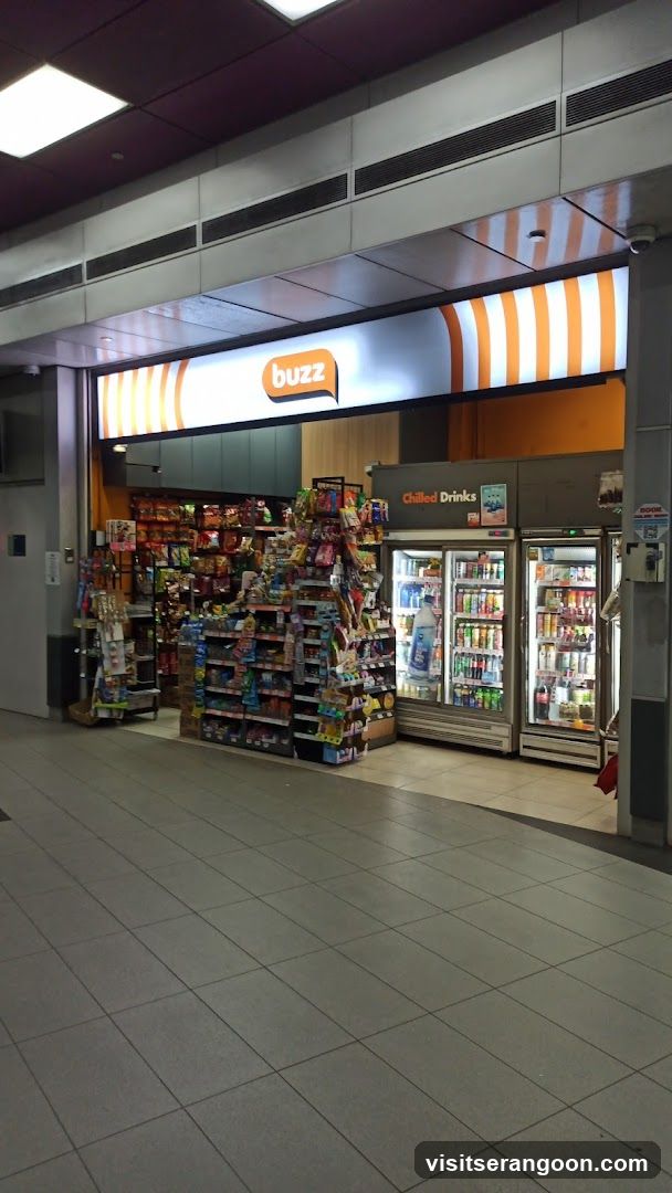 A brightly lit storefront for a convenience store named 'buzz'. The entrance features a prominent sign with orange and white vertical stripes. Inside, shelves are stocked with various snacks and goods, and a refrigerated section labeled 'Chilled Drinks' is visible on the right.