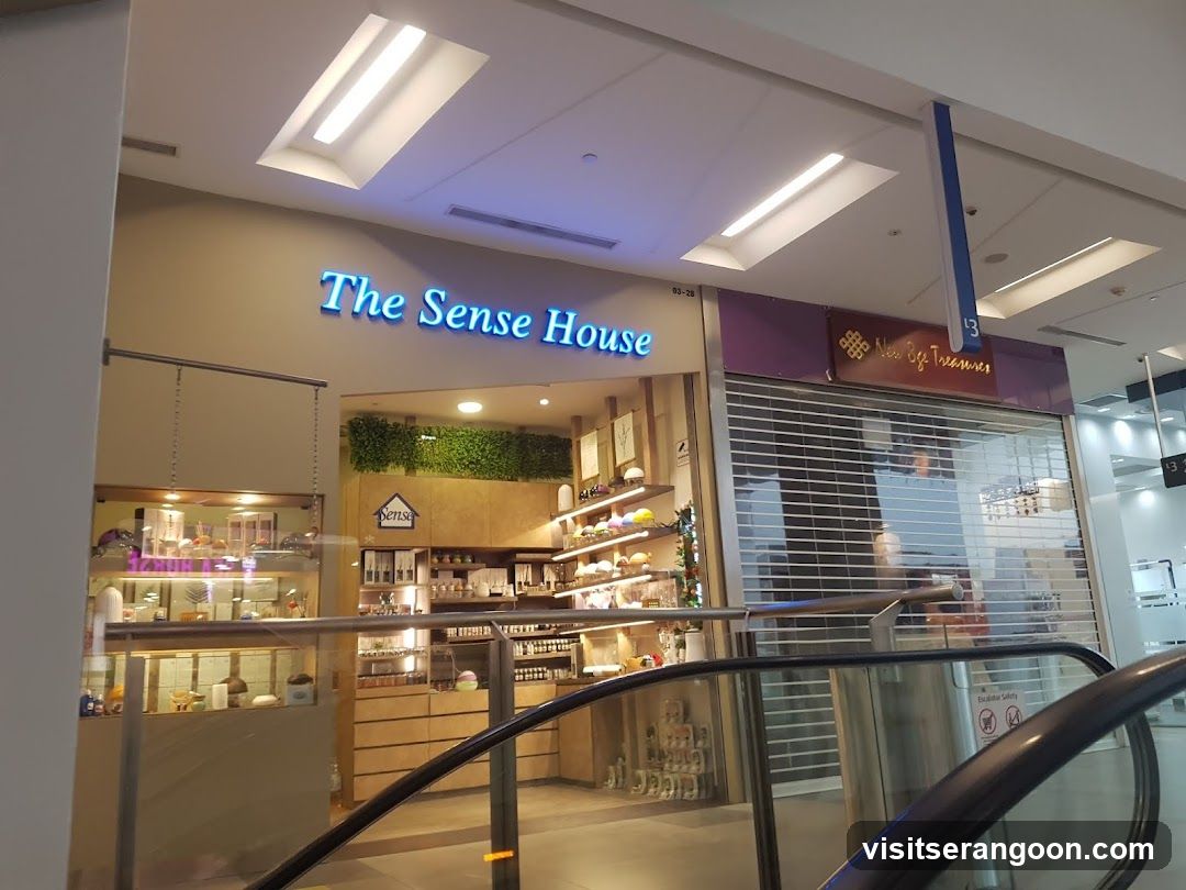 A view of a retail store entrance located inside a shopping mall. The store features a prominent blue neon sign reading 'The Sense House' above the entrance. The interior is visible through large glass windows, displaying shelves stocked with various products, including jars and bottles, and a green moss wall feature. To the right, an adjacent store with a purple sign is partially visible, and a metal security shutter is pulled down over its entrance.