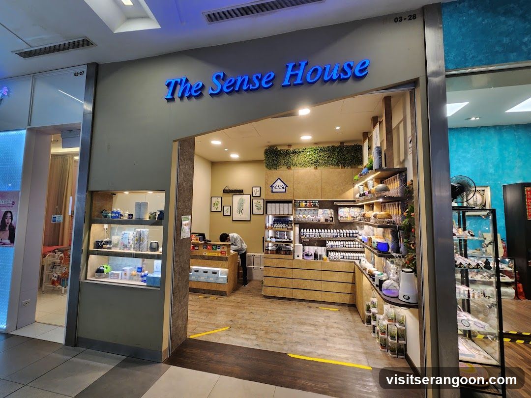 A view of the exterior entrance to a retail store named 'The Sense House'. The store name is displayed in large, blue, illuminated letters above the entrance. The storefront features a wide, open entryway with dark wood trim. Inside, the shop has light wood flooring and walls. A person is visible working behind a wooden counter. To the right of the entrance, there are shelves stocked with various products, including bottles and jars. To the left, a glass display case shows more items. The store number '03-28' is visible in the top right corner of the facade.