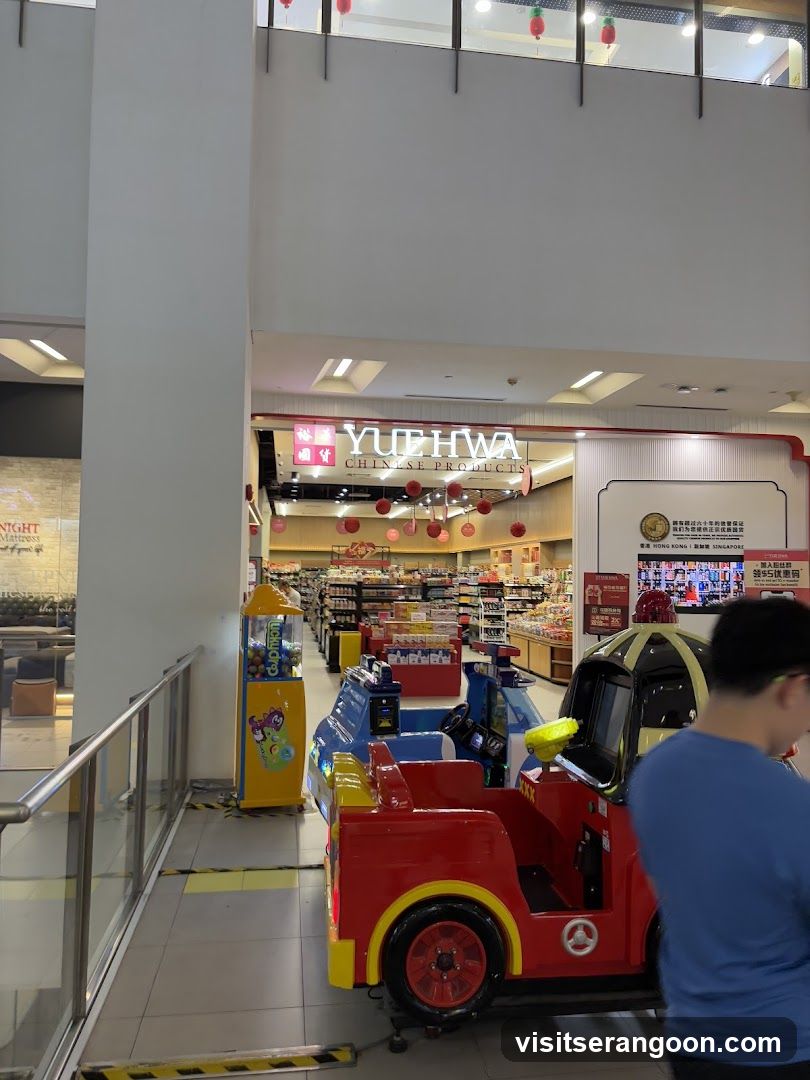 A view of the entrance to a Yue Hwa Chinese Products store located inside a shopping mall. The store features a prominent illuminated sign with the name 'YUE HWA' and 'CHINESE PRODUCTS'. The entrance is decorated with hanging red lanterns. In the foreground, there are several colorful children's arcade ride-on cars, including a red fire truck and a blue vehicle, positioned near a yellow claw machine. A person in a blue shirt is partially visible on the right side.