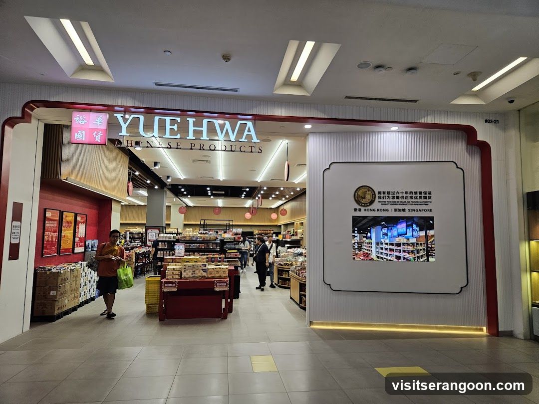 A wide-angle view of the entrance to a Yue Hwa Chinese Products store located inside a shopping mall. The storefront features a large, distinctive red and white frame with the store name 'YUE HWA' and 'CHINESE PRODUCTS' illuminated in white letters above the entrance. Inside, the store is brightly lit with wooden shelving and displays of various goods. A customer carrying a green bag is walking near the entrance, and a large informational display board stands to the right of the doorway.