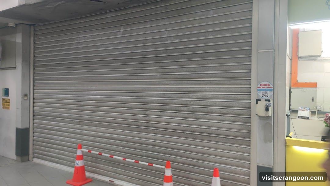 A closed storefront featuring a large, grey metal roller shutter door. The entrance is blocked by orange traffic cones and a red-and-white striped barrier pole. To the right of the shutter, a small sign reading 'BOOK' is visible on the wall next to an electrical box. A yellow counter is partially visible on the far right edge.