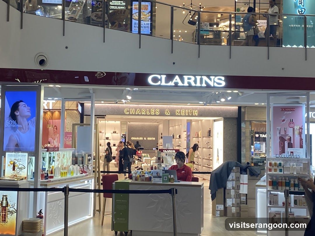 A view of a retail storefront inside a shopping mall. The main sign reads 'CLARINS' in large white letters on a maroon background. Below it, another store entrance is visible with the sign 'CHARLES & KEITH'. The Clarins counter features a staff member in a red shirt, product displays, and promotional posters, including one advertising '50% LIGHTER FEEL'.