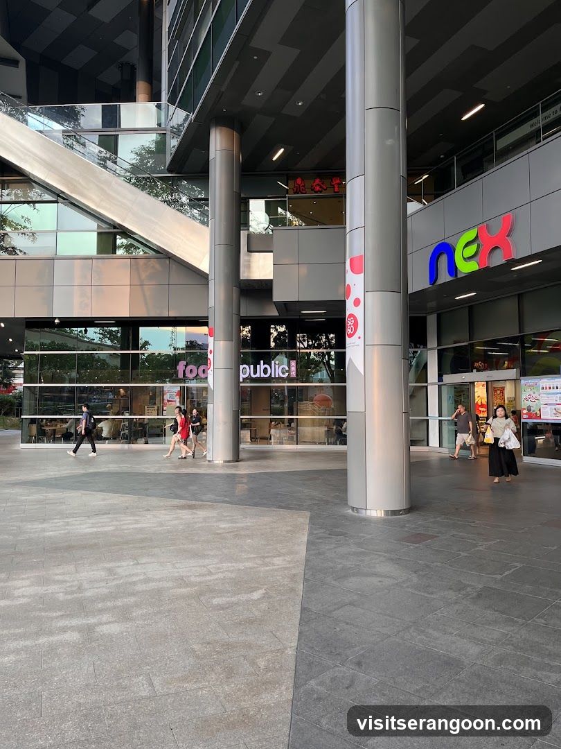 A wide-angle shot of the ground-level exterior of a modern shopping mall. Large silver structural pillars support the upper floors. The mall's name, 'nex', is displayed in colorful letters on the facade. Visible storefronts include 'food public!' and a restaurant with red lanterns. Several pedestrians are walking across the paved plaza.