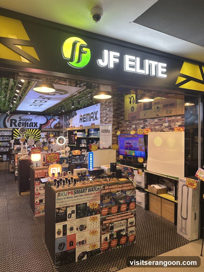 A brightly lit electronics store entrance with a large black sign reading 'JF ELITE' in white letters next to a green and yellow logo. The interior is visible, displaying shelves of gadgets, a television screen, and promotional posters for brands like Remax and Super Remax.