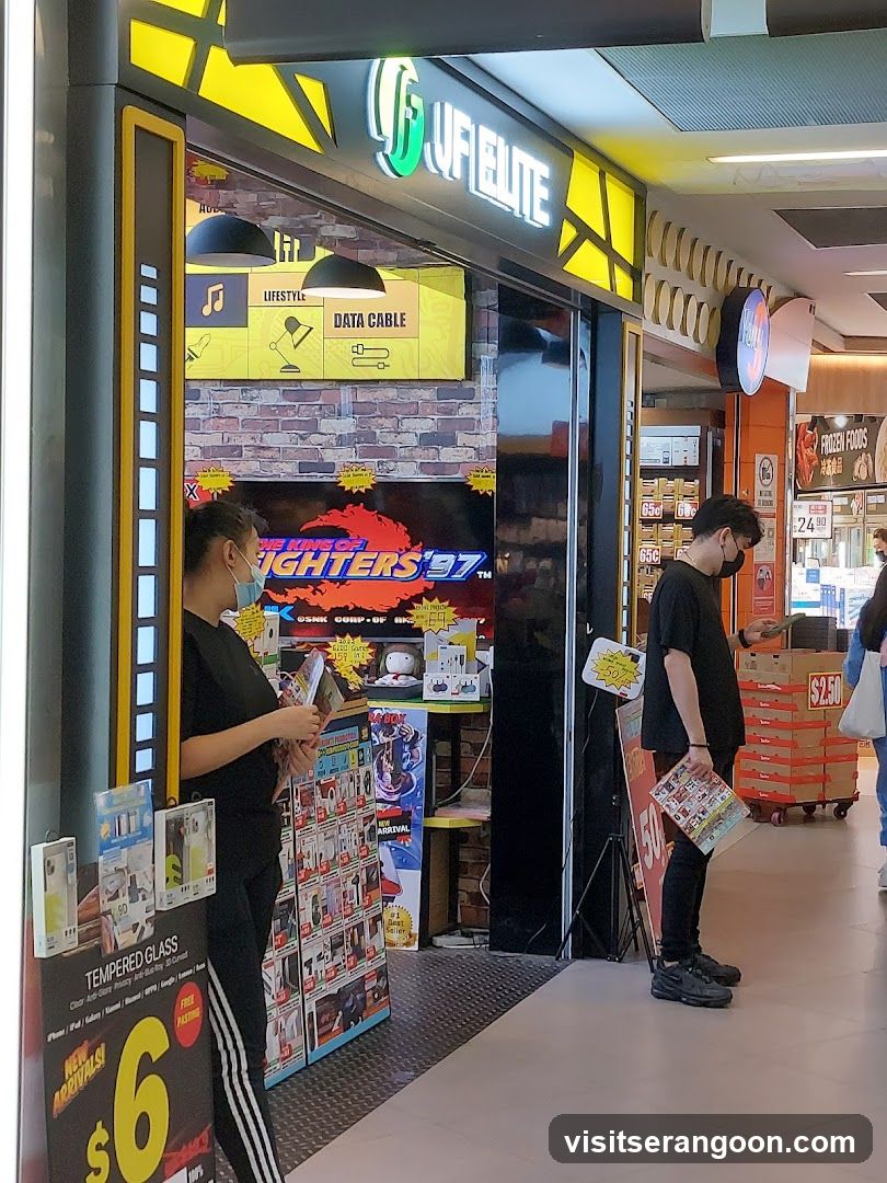 A view of the JF Elite storefront located in a shopping mall. The store features a black and yellow color scheme with a brick-patterned interior wall. A large neon sign reads 'JF ELITE' above the entrance. Inside, a 'FIGHTERS '97' arcade game is visible. Two customers wearing face masks are browsing merchandise, including phone accessories and manga books displayed on racks outside the store.