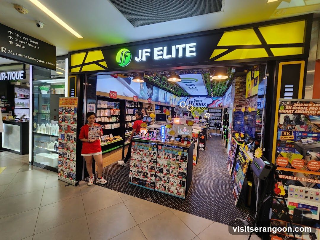 A brightly lit electronics store entrance with a large black and yellow sign reading 'JF ELITE' and a green logo. Two staff members in red shirts stand near the entrance, one holding a product box. The store interior is visible, displaying shelves of boxed merchandise, posters, and promotional banners. A directional sign to the left points to 'The Pet Safari', 'Shaw Theaters', and 'Daiso'.