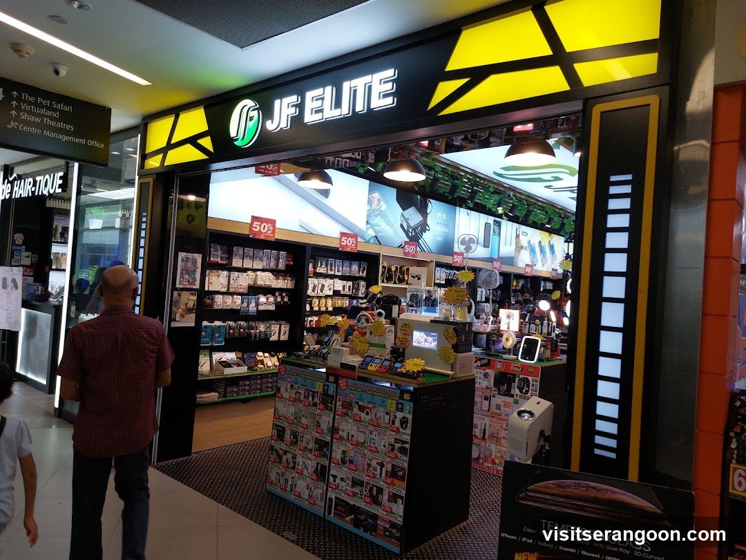 A brightly lit electronics store named 'JF ELITE' with a black and yellow facade. The entrance is open, revealing shelves stocked with mobile accessories, phone cases, and gadgets. A man in a red shirt stands near the entrance, and a sign for 'The Pet Safari' is visible to the left.