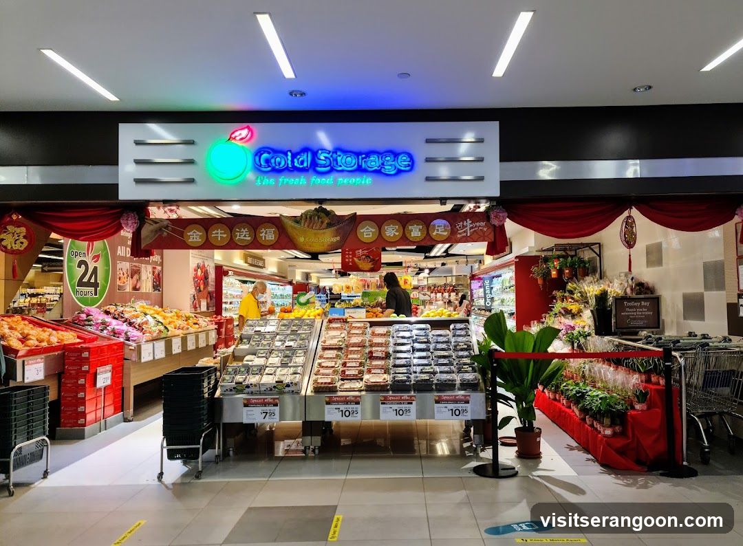 A brightly lit supermarket storefront with a large illuminated sign reading 'Cold Storage' and the slogan 'Live fresh. Feel good.' The entrance is decorated with red banners and hanging ornaments, suggesting a festive season. Inside, aisles are stocked with fresh produce, packaged goods, and refrigerated items. Staff and customers are visible near the checkout and display areas.