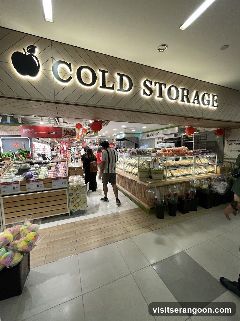 A brightly lit entrance to a Cold Storage supermarket. The store name is displayed in large, illuminated letters on a geometric-patterned facade above the entrance. Inside, customers are browsing aisles filled with fresh produce, baked goods, and flowers. Red lanterns hang from the ceiling, suggesting a festive season.