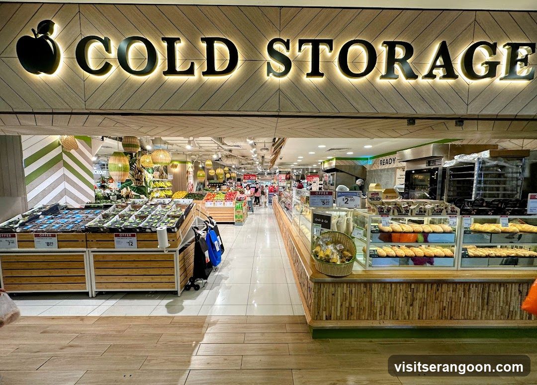 A brightly lit supermarket entrance featuring a large, illuminated sign reading 'COLD STORAGE' with a black apple logo. The interior is visible, showing aisles of fresh produce on wooden displays to the left and a deli counter with baked goods to the right. The flooring is light-colored tile, and the ceiling has modern lighting fixtures.