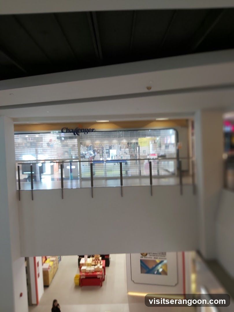 A view of a storefront located on an upper level of a shopping mall. The store has a large glass facade with horizontal blinds partially drawn. A sign reading 'Challenger' is visible above the entrance. A metal railing runs along the walkway in front of the store.