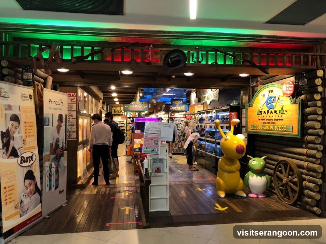 A brightly lit souvenir shop entrance featuring a rustic log cabin facade. A large wooden sign on the right reads 'The SAFARI SINGAPORE' and 'Paw-More Gifts'. The interior is filled with shelves of merchandise, and large cartoon character statues (a yellow creature and a green penguin) stand near the entrance. Several customers are browsing inside.