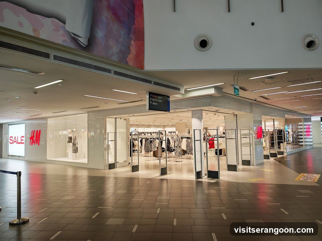 A wide shot of an H&M store entrance located within a shopping mall. The storefront features large glass windows and doors, with a prominent red 'SALE' sign and the H&M logo displayed on the left. Inside, clothing racks are visible. The mall corridor has tiled flooring and overhead lighting.