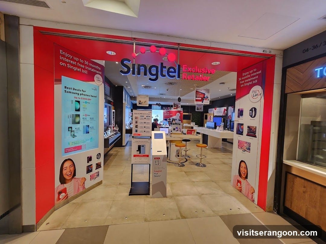 A view of the entrance to a Singtel Exclusive Retailer store located within a shopping mall. The storefront features a large red facade with the blue 'Singtel' logo and 'Exclusive Retailer' text above the entrance. Large posters on either side of the entrance advertise promotions, including 'Enjoy up to 36 months Interest Free Installments' and 'Best Deals for Samsung phones here!'. Inside, the store is brightly lit with white counters, display shelves, and seating areas.