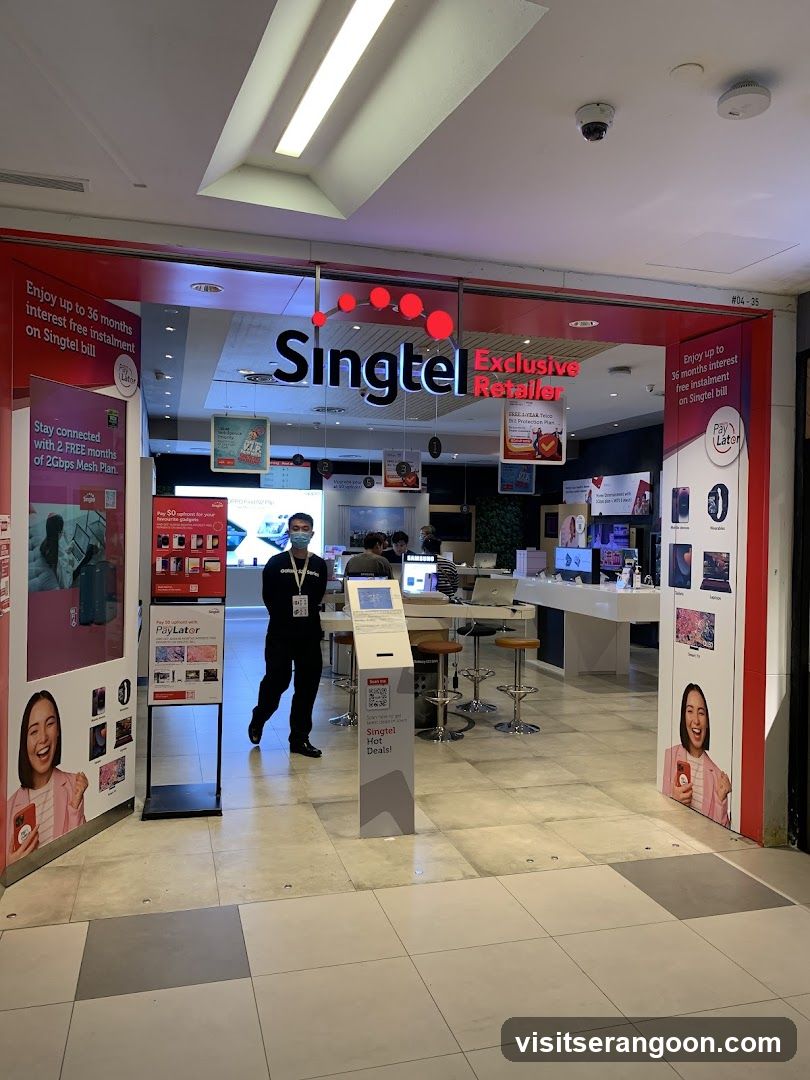 A brightly lit storefront with a large red and white sign reading 'Singtel Exclusive Retailer'. The entrance is open, revealing a modern interior with white counters, stools, and digital displays. A staff member wearing a mask stands near the entrance. Promotional posters on the pillars advertise interest-free installments and free months of Galaxy Mesh Plan.