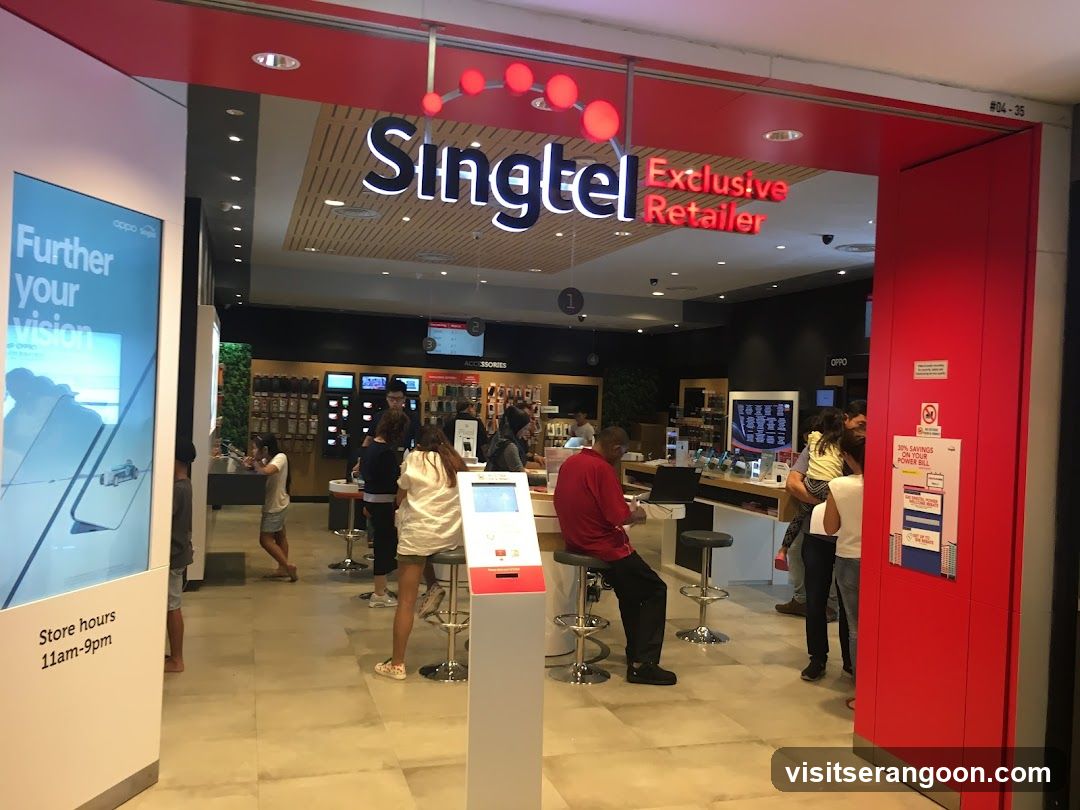 A brightly lit storefront with a large red overhead sign reading 'Singtel Exclusive Retailer'. The entrance is open, revealing a modern interior with wooden ceilings and tiled floors. Inside, customers are browsing products and interacting with staff at service counters. A digital kiosk stands near the entrance, and a poster on the left wall advertises 'Further your vision'.