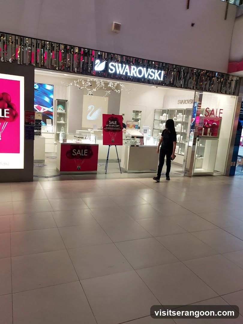 A brightly lit Swarovski jewelry store located within a shopping mall. The storefront features a prominent illuminated sign with the brand name and swan logo. The entrance is open, revealing white display counters, glass cabinets, and hanging crystal chandeliers. Large pink sale signs are visible inside and outside the store, and a customer is browsing near the entrance.