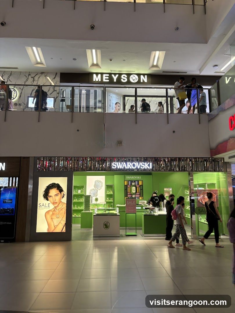 A brightly lit Swarovski store located on the ground floor of a multi-level shopping mall. The storefront features a distinctive green color scheme with glass display cases and a large illuminated sign displaying the brand name. A large advertising poster showing a model is visible to the left of the entrance. Several people are walking past the store, and other shoppers can be seen on the upper balcony level above.