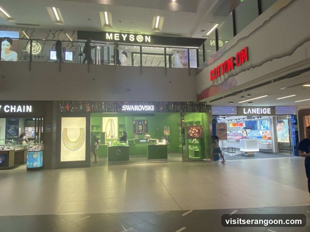 A view of a shopping mall corridor showing multiple retail stores on two levels. Visible storefronts include Meyson, Swarovski, and Laneige. Large red signage for Cotton On is also visible on the upper level.
