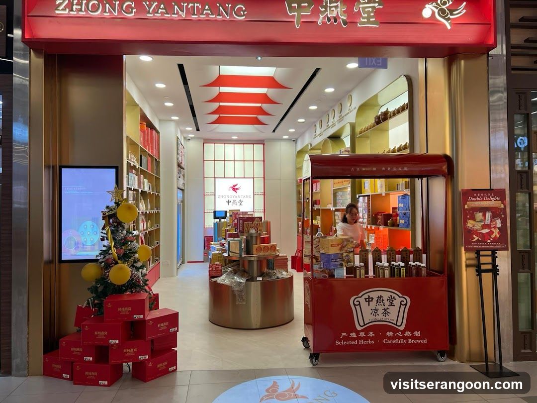 A view of the entrance to a Zhong Yantang store. The storefront features a large red sign with the name 'ZHONG YANTANG' in English and Chinese characters. Inside, the store is brightly lit with gold and red decor, including a distinctive red pagoda-style ceiling fixture. A red mobile cart displaying bottled products stands near the entrance, and a small Christmas tree with red gift boxes is visible on the left.