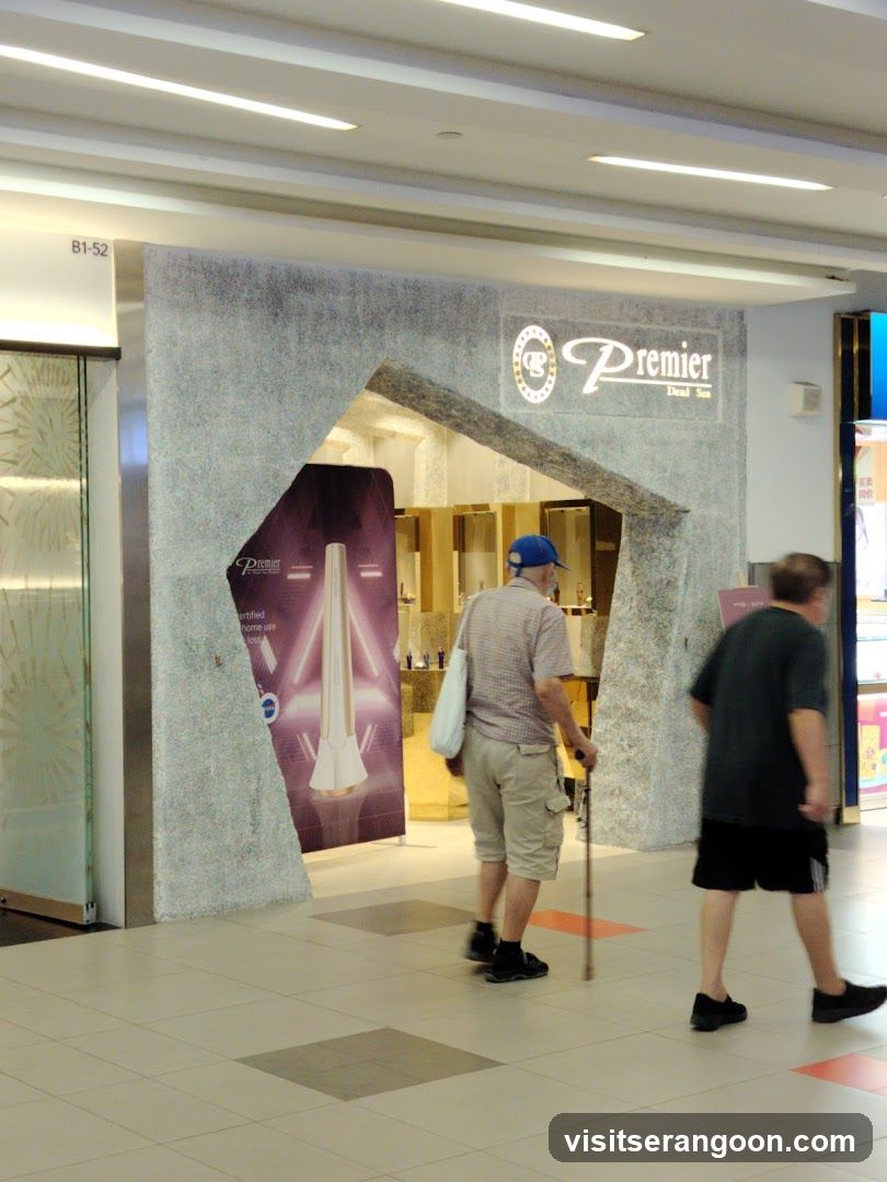 A view of a store entrance inside a shopping mall. The storefront features a textured, grey stone facade with an arched entryway. A sign above the entrance reads 'Premier' next to a circular logo. Inside, gold-toned shelving and a large purple poster advertising a product are visible. Two men are walking past the store in the foreground.