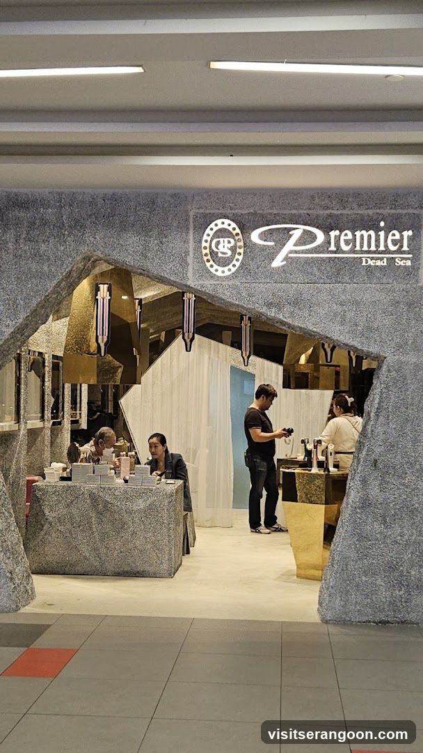 A view of a retail kiosk or store entrance designed to look like a stone cave. The facade features a rough, grey, textured material forming an arched entryway. Above the entrance, a sign displays the brand name 'Premier' in white script alongside a circular logo and the text 'Dead Sea'. Inside, several people are visible, including staff behind counters and customers browsing products.