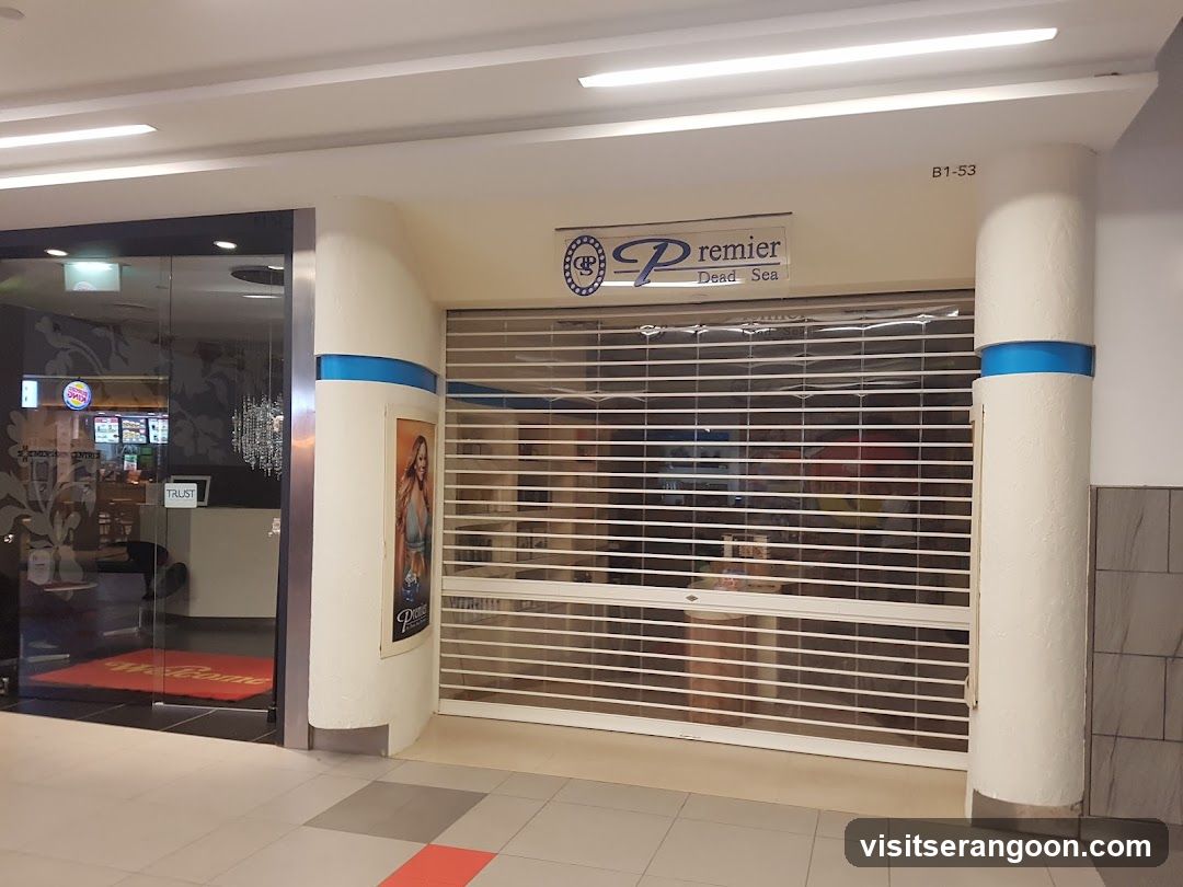 A closed storefront with a white rolling metal gate. A sign above reads 'Premier Dead Sea'. The entrance is flanked by two white pillars with blue stripes. To the left, a glass door reveals a glimpse of an interior with a red floor mat.