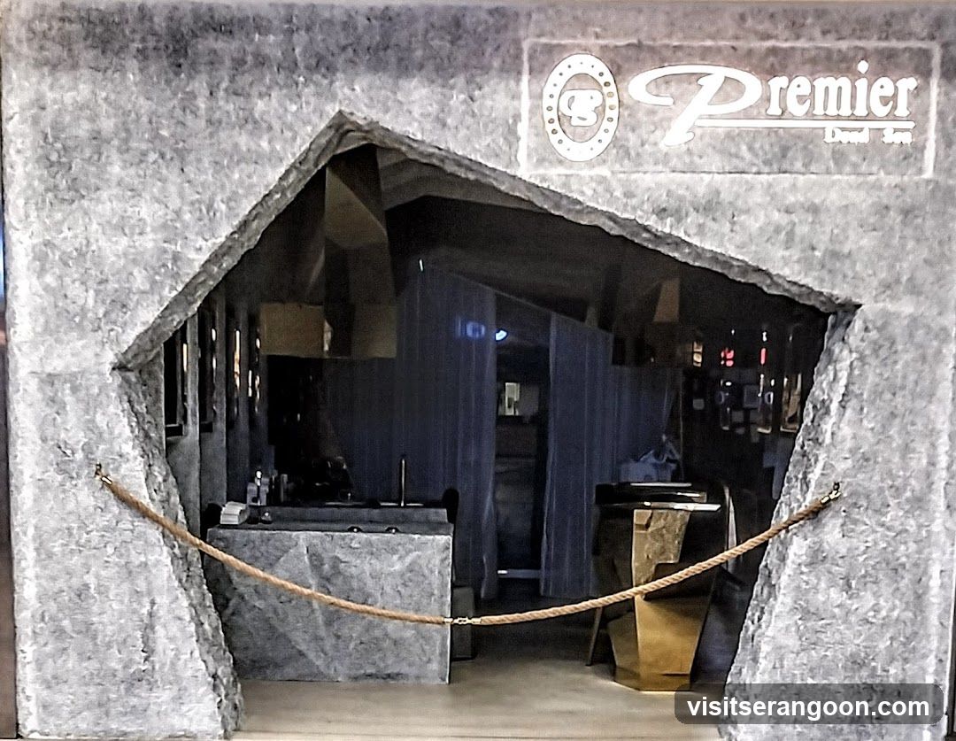A view through a large, jagged hole in a grey, textured wall that resembles cracked stone or concrete. Inside the opening, a salon interior is visible with dark counters, mirrors, and blue curtains. A sign reading 'Premier' is mounted on the wall above the opening.