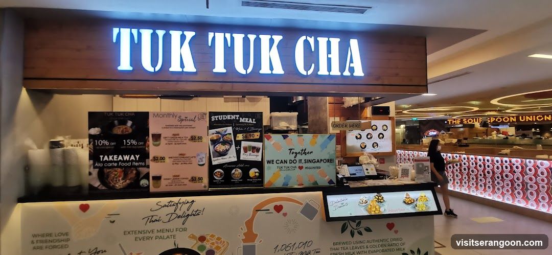 A brightly lit storefront for a beverage shop named 'TUK TUK CHA' with large white illuminated letters on a dark wooden sign. The counter displays several promotional posters, including offers for 10% and 15% off takeaway items, student meal deals, and a 'Together We Can Do It, Singapore!' campaign. The interior features a long counter with a distinctive red and white patterned front, and another business, 'The Soup Spoon Union', is visible in the background.