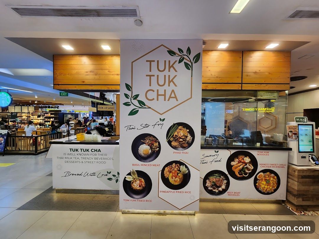 A brightly lit food stall in a mall with a large central banner displaying the name 'TUK TUK CHA' and images of various Thai dishes. The stall features a wooden counter, a glass partition, and a digital ordering kiosk on the right. A smaller sign on the left counter describes the brand's offerings.
