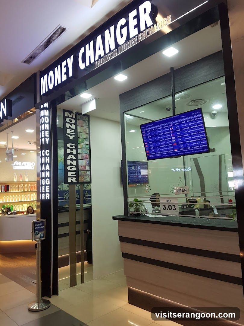 A brightly lit storefront for a money exchange business located inside a shopping mall. The shop features a large black sign with white lettering reading 'MONEY CHANGER' and 'THOMSON MONEY EXCHANGE'. A digital screen displaying currency exchange rates is visible through the glass window, along with a counter and an 'OPEN' sign.