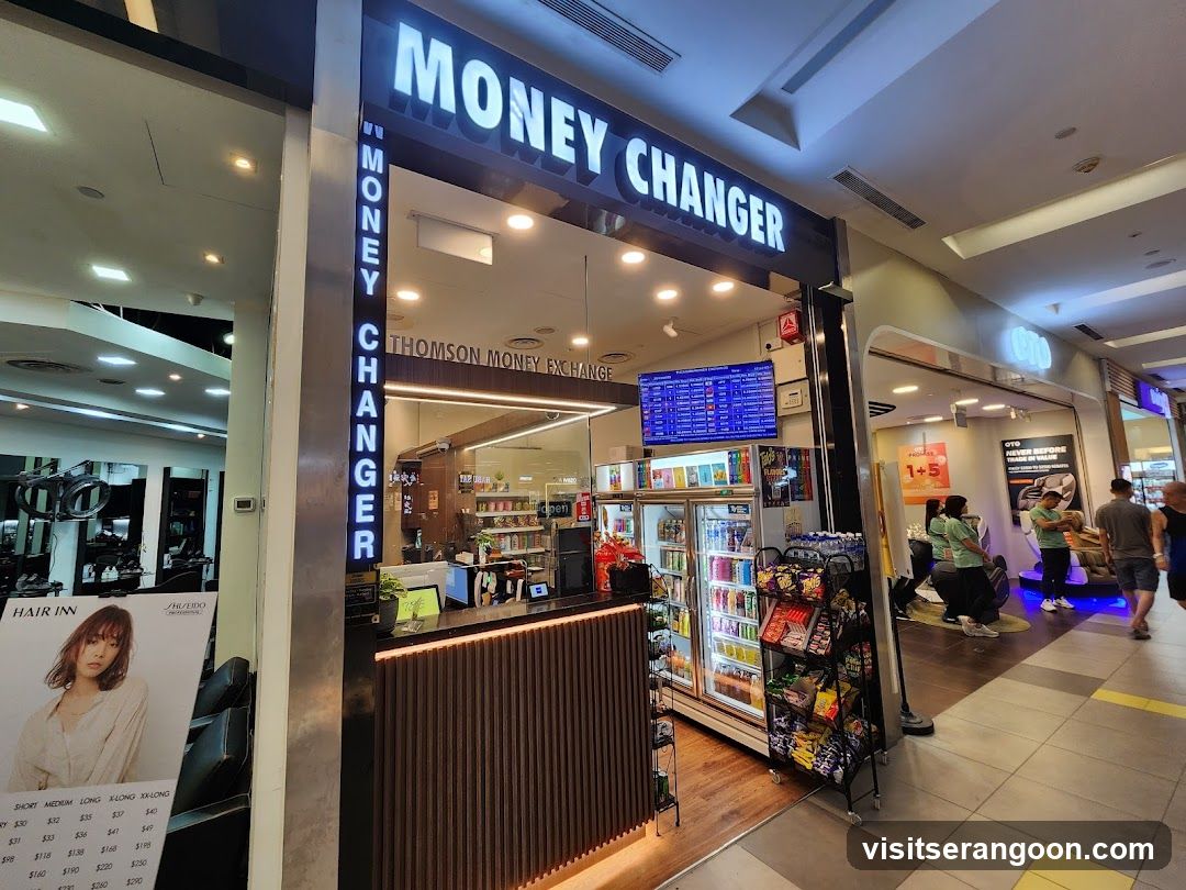 A brightly lit storefront for a money exchange located inside a shopping mall. The business features a large overhead sign reading 'MONEY CHANGER' in white letters, with 'THOMSON MONEY EXCHANGE' written below. A vertical neon sign on the left pillar also reads 'MONEY CHANGER'. The interior is visible through the open entrance, showing a wooden counter, a digital currency rate display screen, and refrigerated beverage coolers stocked with drinks.