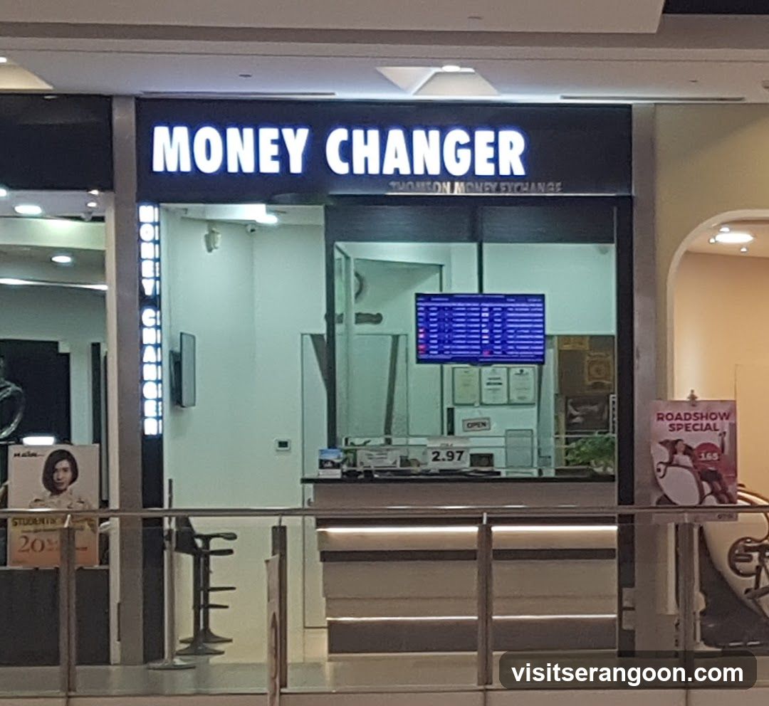 A storefront with a large illuminated sign reading 'MONEY CHANGER' above the entrance. The shop has glass walls and doors, revealing an interior with a counter, a digital screen displaying exchange rates, and an 'OPEN' sign. A vertical sign on the left also reads 'MONEY CHANGER'.
