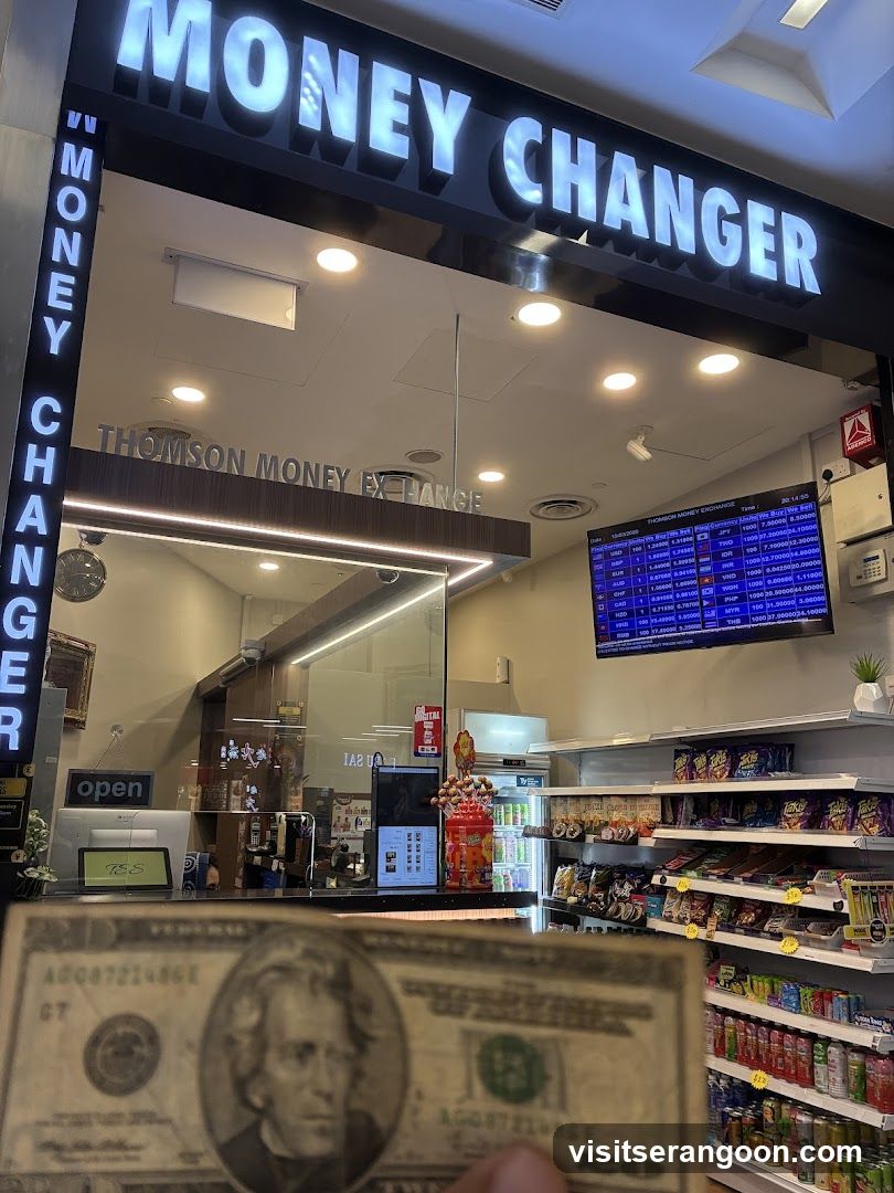A brightly lit storefront with a large, illuminated sign reading 'MONEY CHANGER' in white letters on a black background. A vertical sign on the left also reads 'MONEY CHANGER'. The interior is visible through a glass partition, showing a counter, a digital screen displaying exchange rates, and shelves stocked with various snacks and drinks. A hand holding a US twenty-dollar bill is in the foreground.