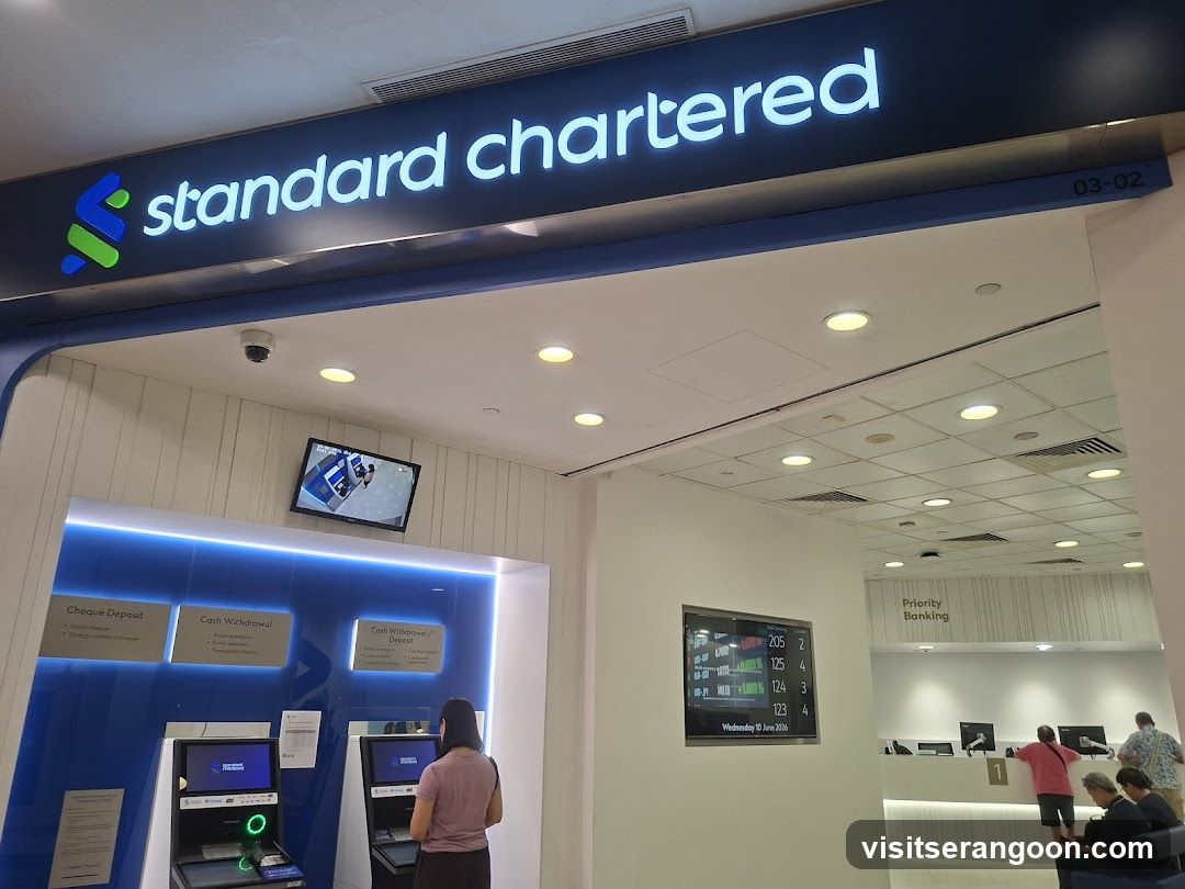 A view of the entrance to a Standard Chartered bank branch. The illuminated blue sign with the company name and logo is mounted above the entrance. Inside, customers are using ATMs and visiting the Priority Banking counter.